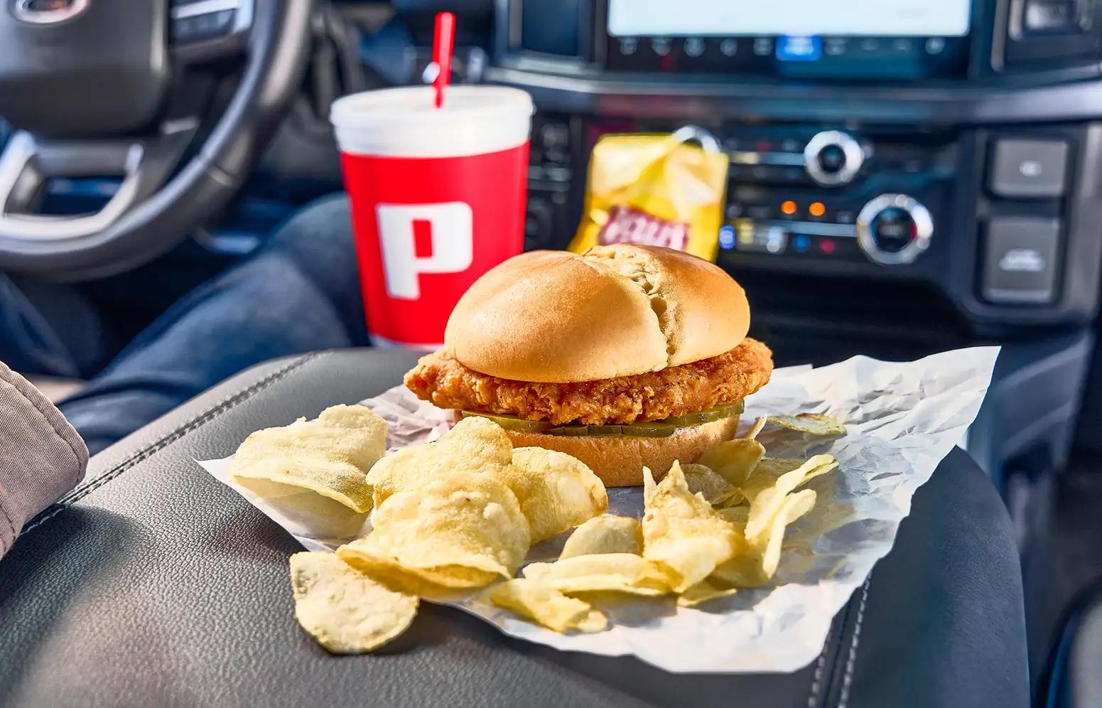 a cripsy chicken sandwich with pickles, a bag of Lay's potato chips, and a large Pilot fountain drink in a car