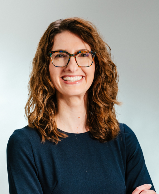 Eva Rigamonti, a woman with curly auburn hair and colorful tortoiseshell glasses, smiling with arms crossed, wearing a navy blue top
