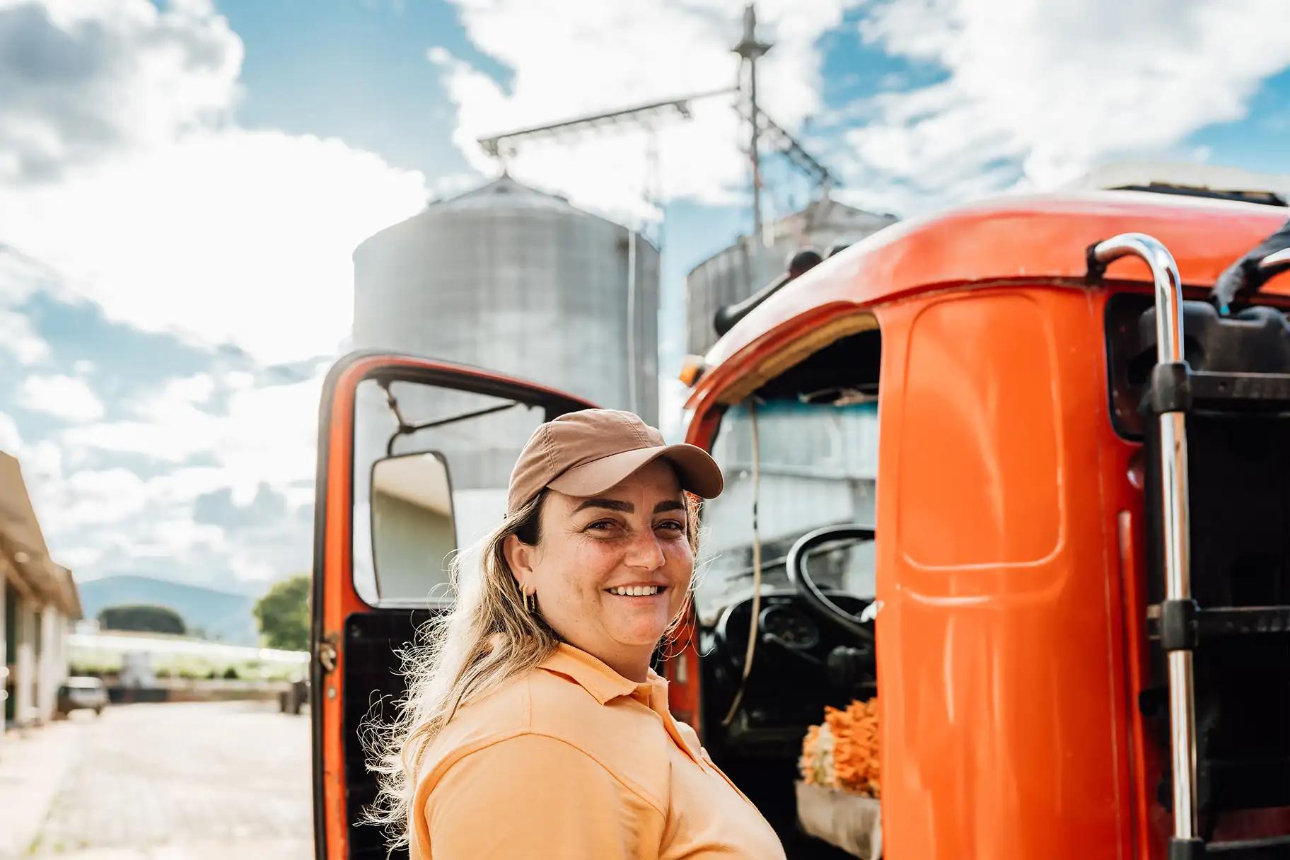Smiling female truck driver standing beside the open door of a red truck cab at a farm facility with grain silos in the background