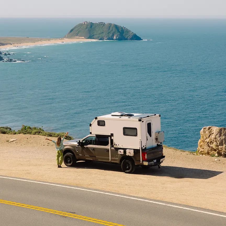A pickup truck with a white camper shell parked beside a coastal road, overlooking blue ocean water and a small rocky island in the distance