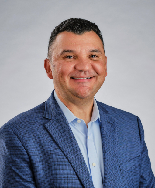Professional headshot of Jesus Guerra wearing a blue blazer and light blue dress shirt, smiling against a gray background