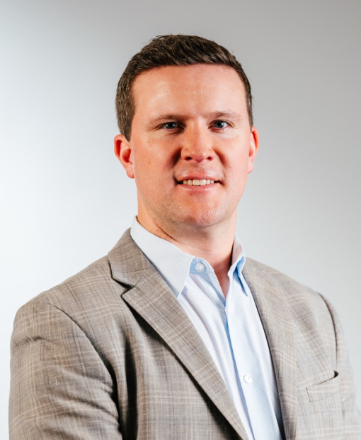 Professional headshot of a man wearing a light gray blazer and light blue dress shirt, smiling, against a neutral gray background