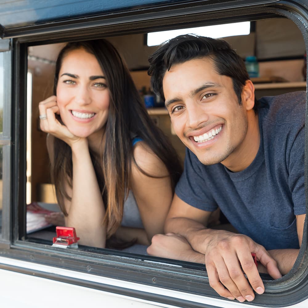 A young couple smiling and looking directly at the camera while peering out of the window of their RV