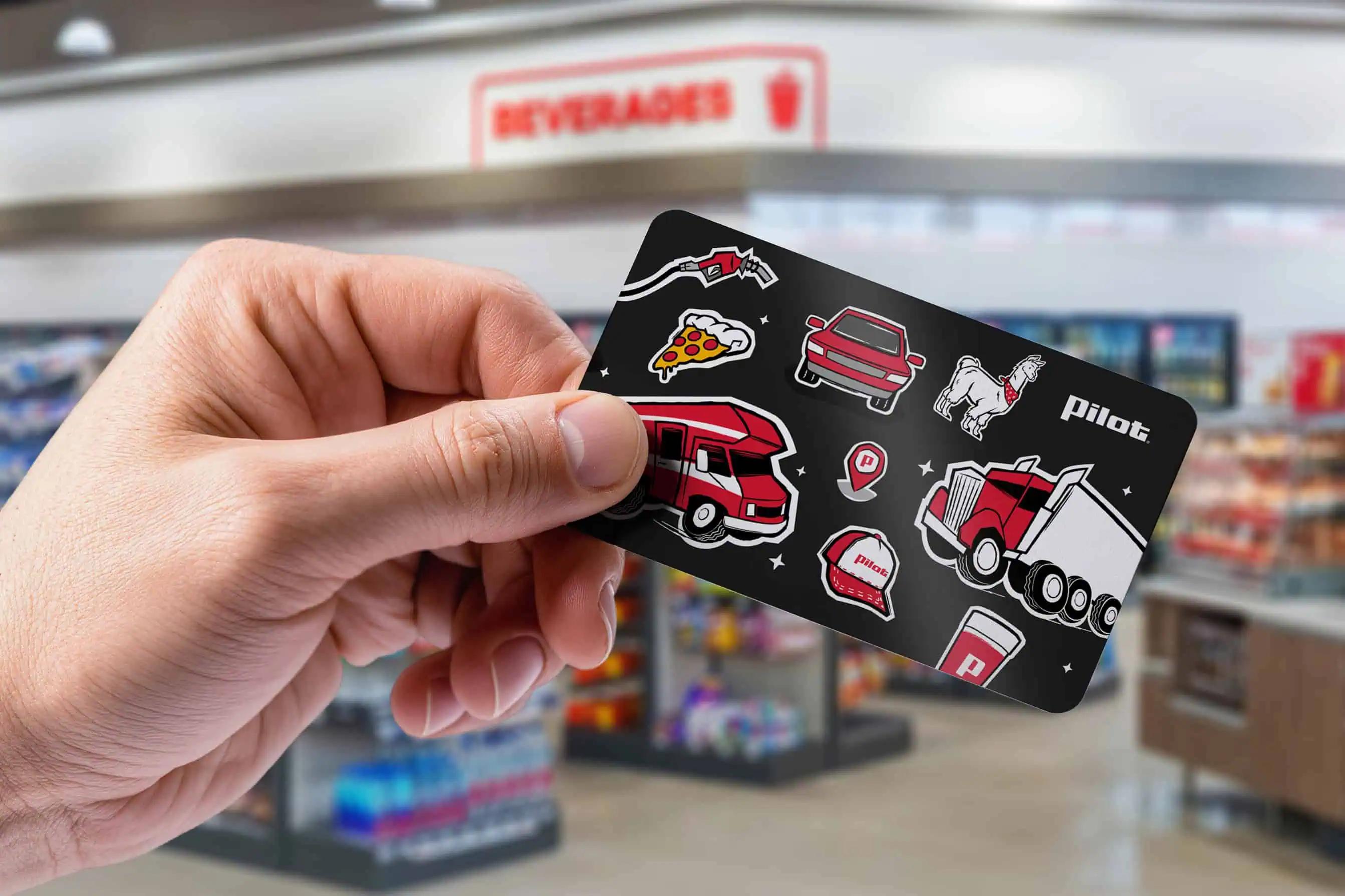 Hand holding a Pilot gift card decorated with illustrations of a semi-truck, RV, fuel nozzle, pizza, coffee cup, and Pilot logo inside a Pilot travel center convenience store.