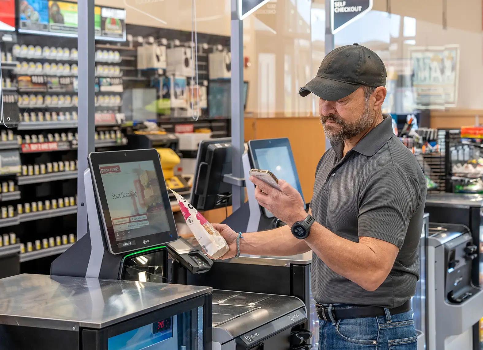 A man in a gray polo shirt and dark cap uses his smartphone at a Pilot Flying J self-checkout kiosk while scanning a packaged food item