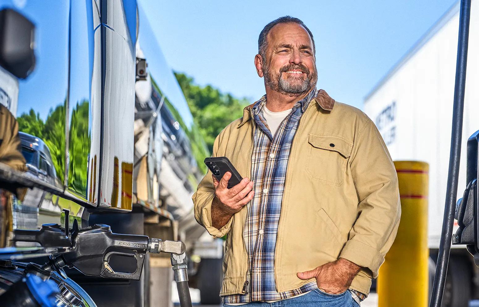 A truck driver stands beside a fuel pump and a large semi-truck, holding a smartphone in one hand while wearing a tan jacket and plaid shirt. The scene takes place outdoors at a fueling station, with another truck and equipment visible in the background.