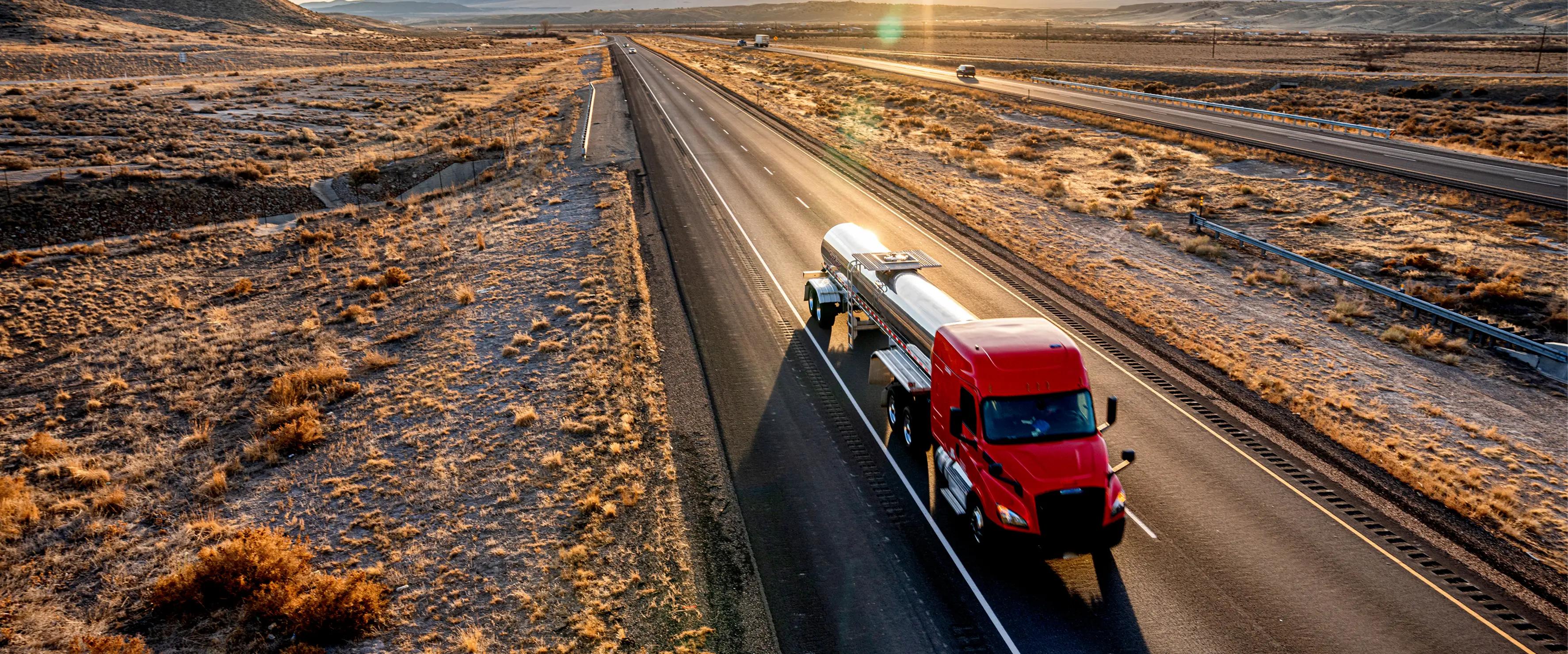 Aerial view of a red semi-truck pulling a silver fuel tanker trailer down a multi-lane highway through an arid desert landscape at sunset