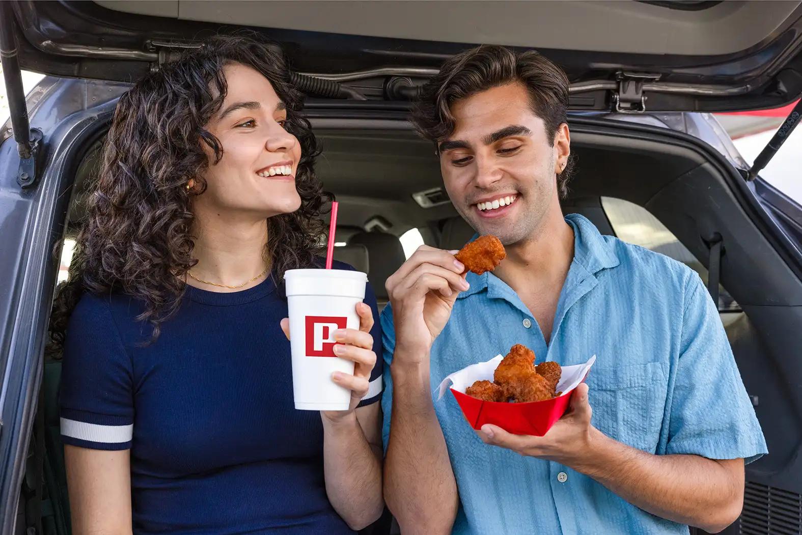 Two smiling people sitting in the open trunk of a car, sharing a basket of fried chicken pieces and a Pilot branded fountain drink with a red straw