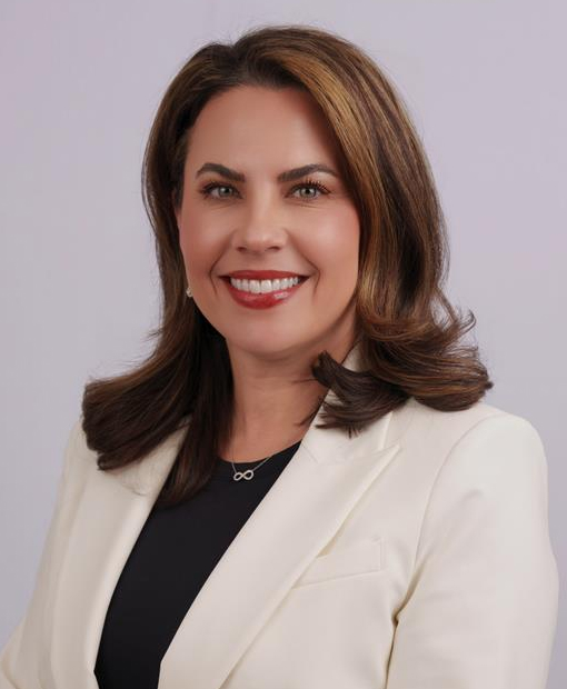 Professional headshot of Kari Irons smiling, with shoulder-length brown hair, wearing a cream blazer over a black top, and a delicate infinity symbol necklace, against a light gray studio background