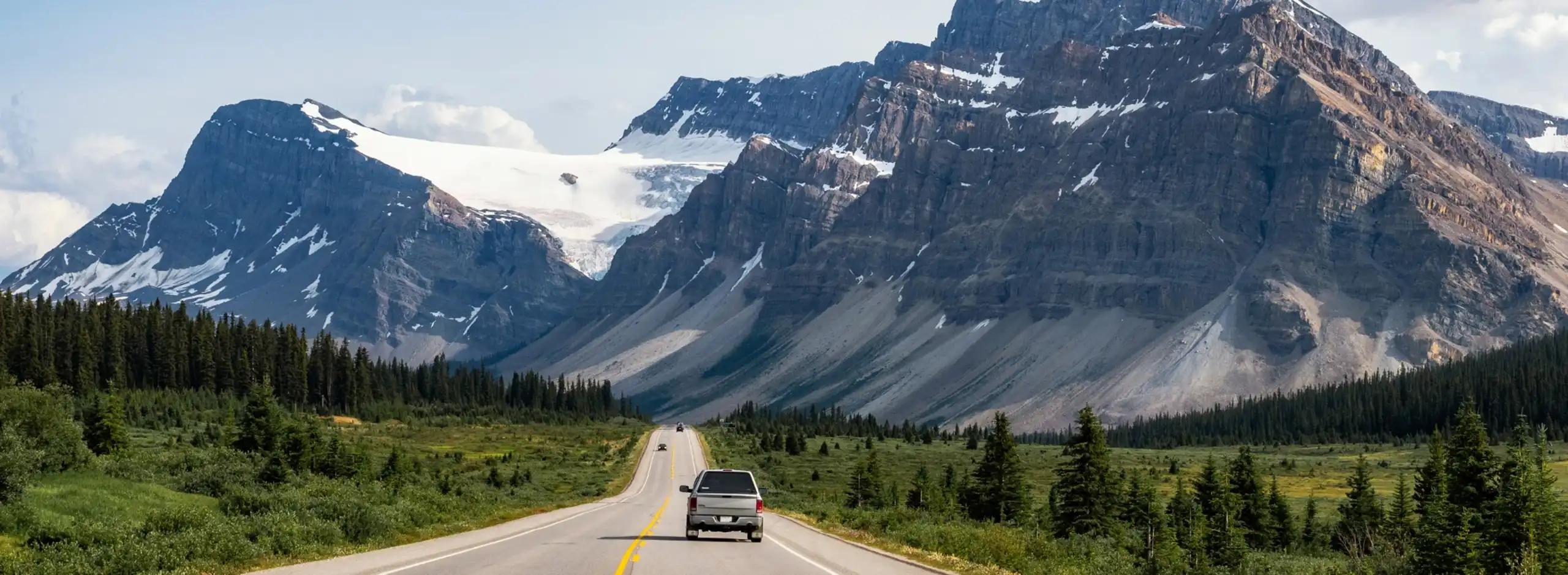 A silver SUV and other vehicles driving along a straight two-lane highway through a lush evergreen valley toward dramatic snow-capped Rocky Mountain peaks with a visible glacier
