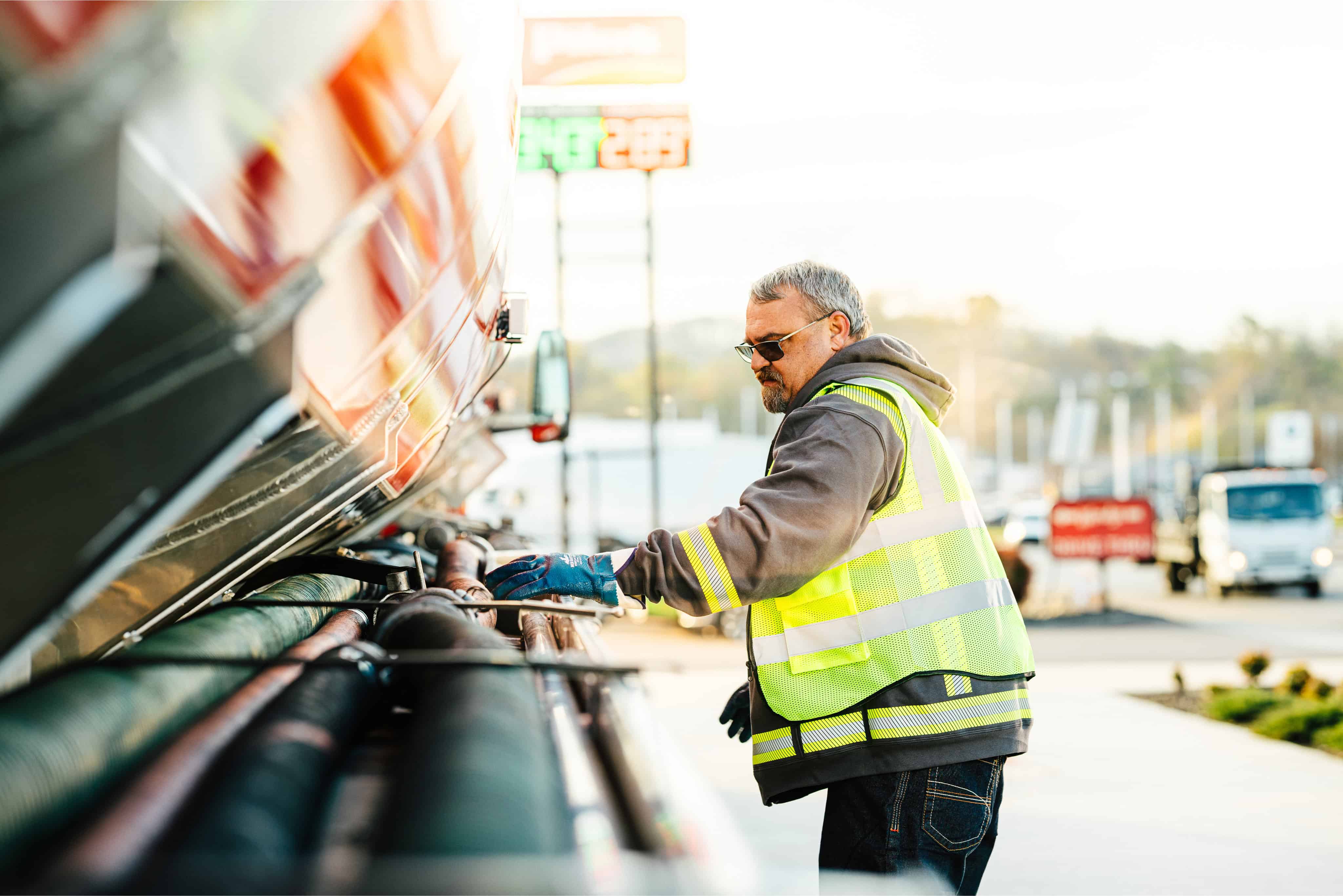 A male worker wearing a yellow high-visibility safety vest, sunglasses, and blue work gloves inspects or connects hoses and fittings along the side of a large tanker truck at a truck stop, with a fuel price sign and other trucks visible in the background
