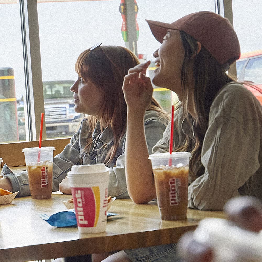 Two young women sitting at a table inside a Pilot Travel Center, each with a Pilot-branded iced coffee drink with a red straw; one woman wears a baseball cap and is eating a snack while looking upward