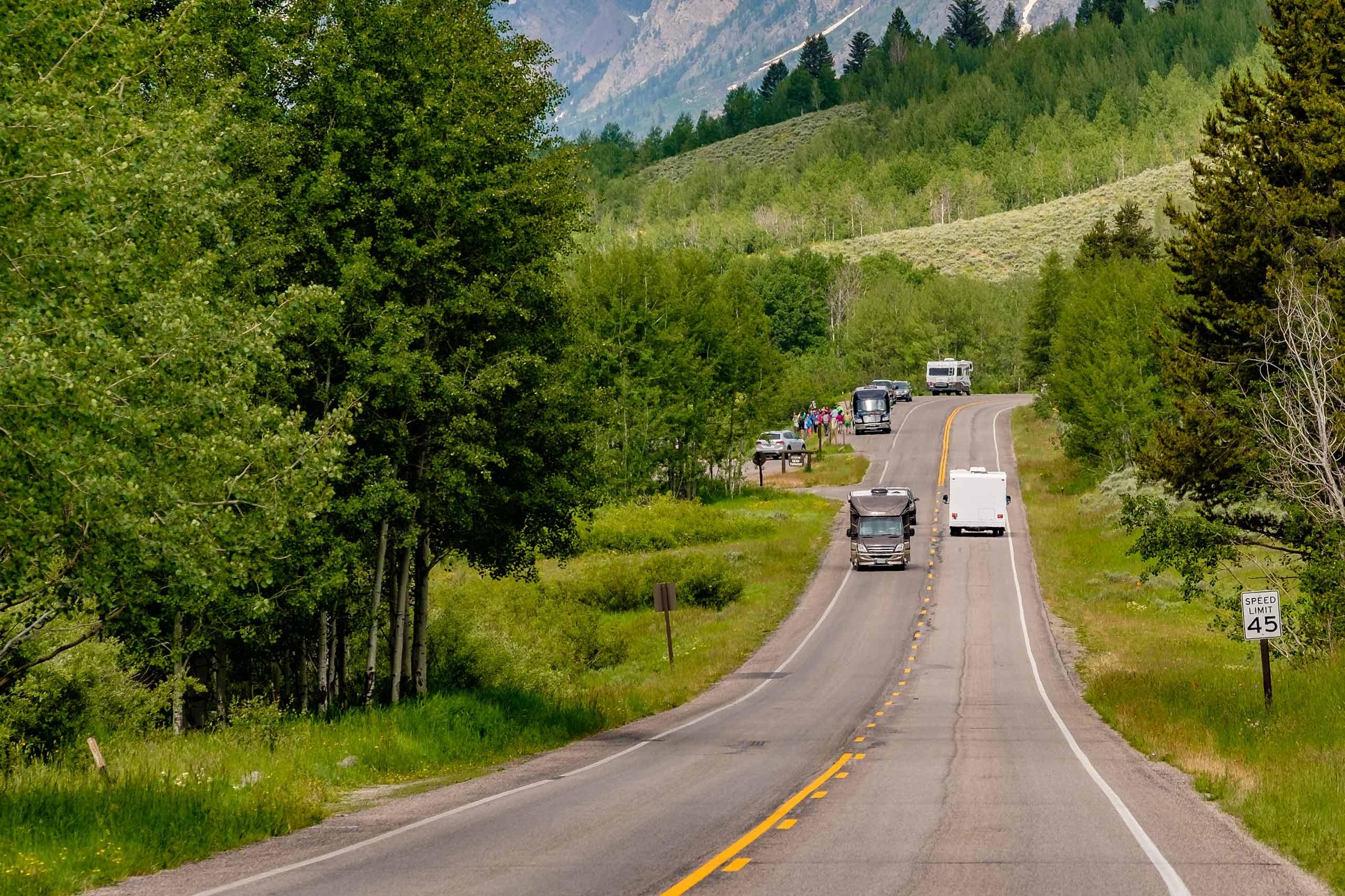 A two-lane mountain highway with a 45 mph speed limit sign, RVs and large vehicles traveling in both directions, lush green trees and meadows on either side, a group of people gathered roadside, and a forested mountain with snow patches in the background