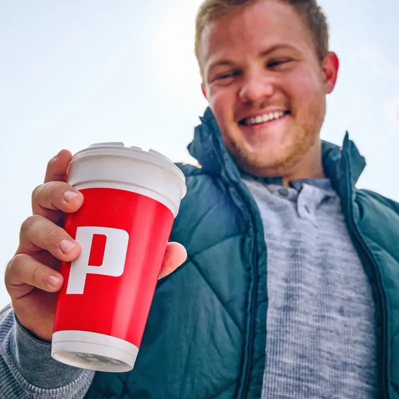 Smiling young man holding a red Pilot coffee cup with a white lid toward the camera