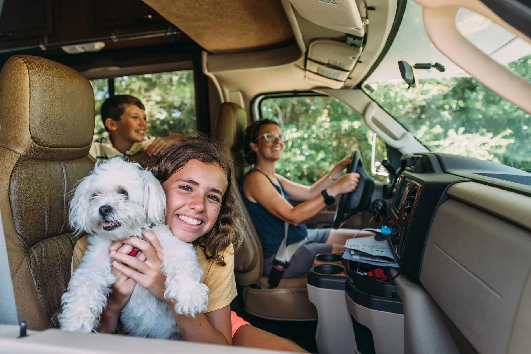 Smiling girl holding a small white fluffy dog in the front cab of an RV, with a woman driving and a boy in the back seat, trees visible through the windshield