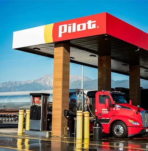 Red semi-truck fueling at a Pilot Travel Center fuel pump, with mountains visible in the background