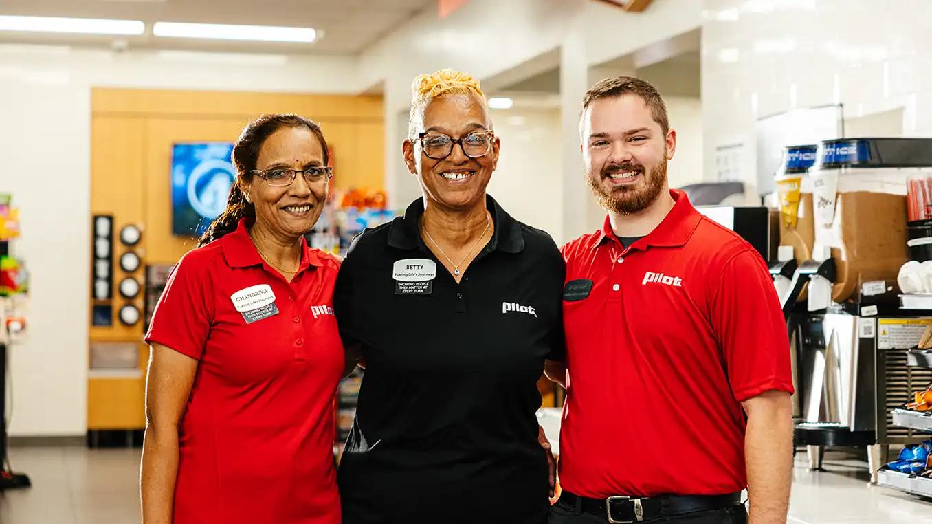 Three smiling Pilot Flying J team members wearing name tags posing together inside a Pilot travel center near the beverage counter