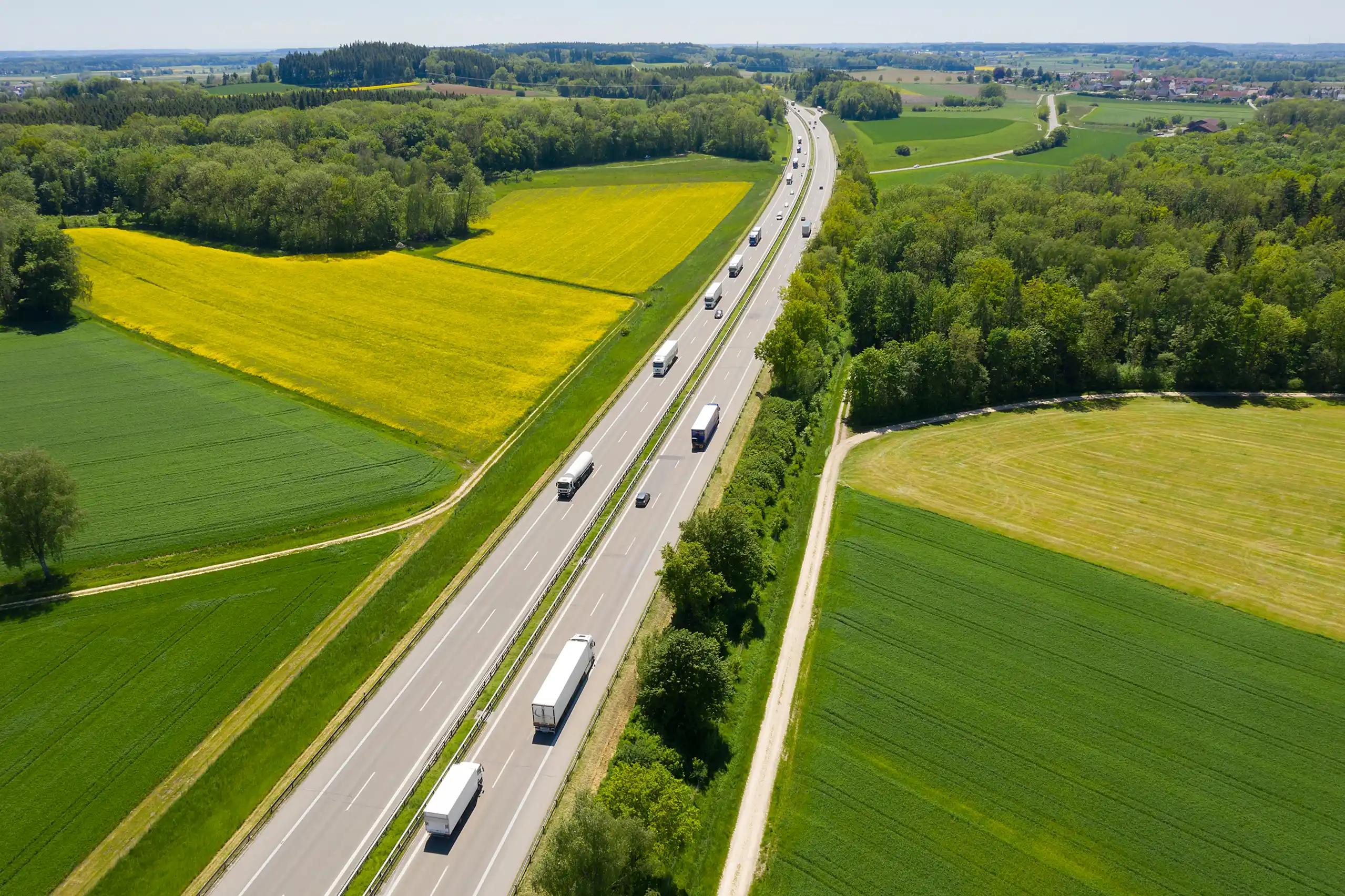 Aerial drone view of a two-lane highway through rural farmland with yellow rapeseed fields, with multiple white semi-trucks and tractor-trailers traveling in both directions