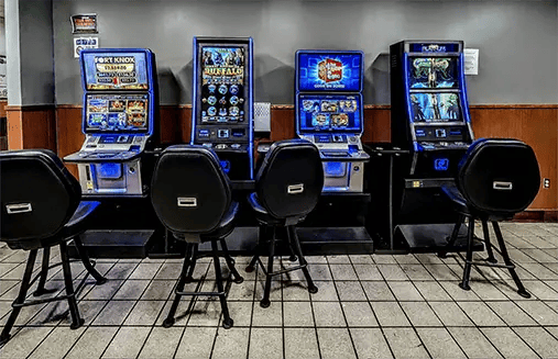 Four slot machines with blue LED lighting, including Buffalo and Fort Knox titles, each with a black bar-height chair, arranged in a row against a gray wall on a tile floor
