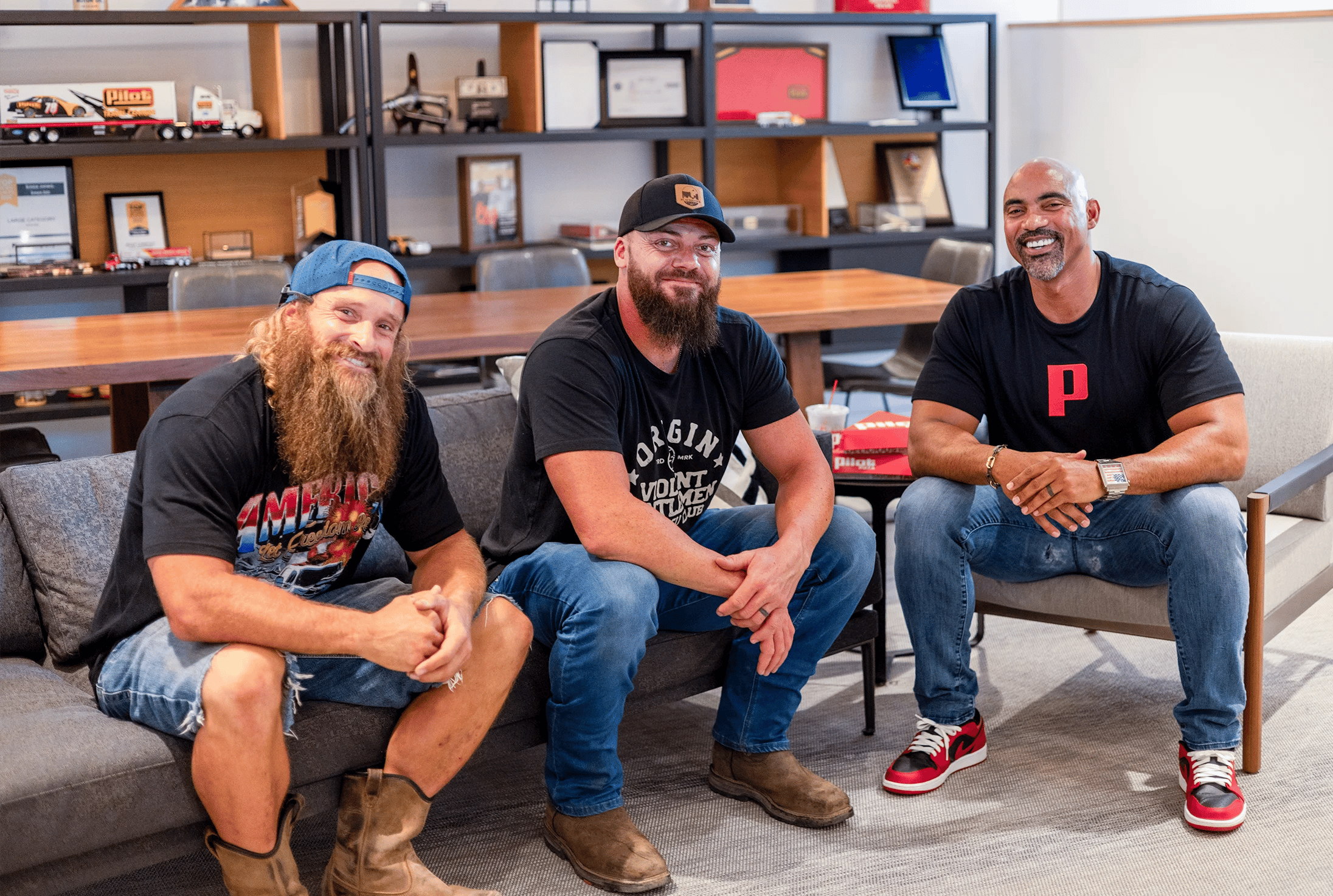 Three men (The Diesel Brothers and CEO Adam Wright) sitting together and smiling in an office setting with shelving displaying model trucks and awards in the background; two men on the left have beards and wear black t-shirts and baseball caps, while the man on the right is bald, wearing a black t-shirt with a red Pilot 'P' logo and red-and-black Air Jordan sneakers