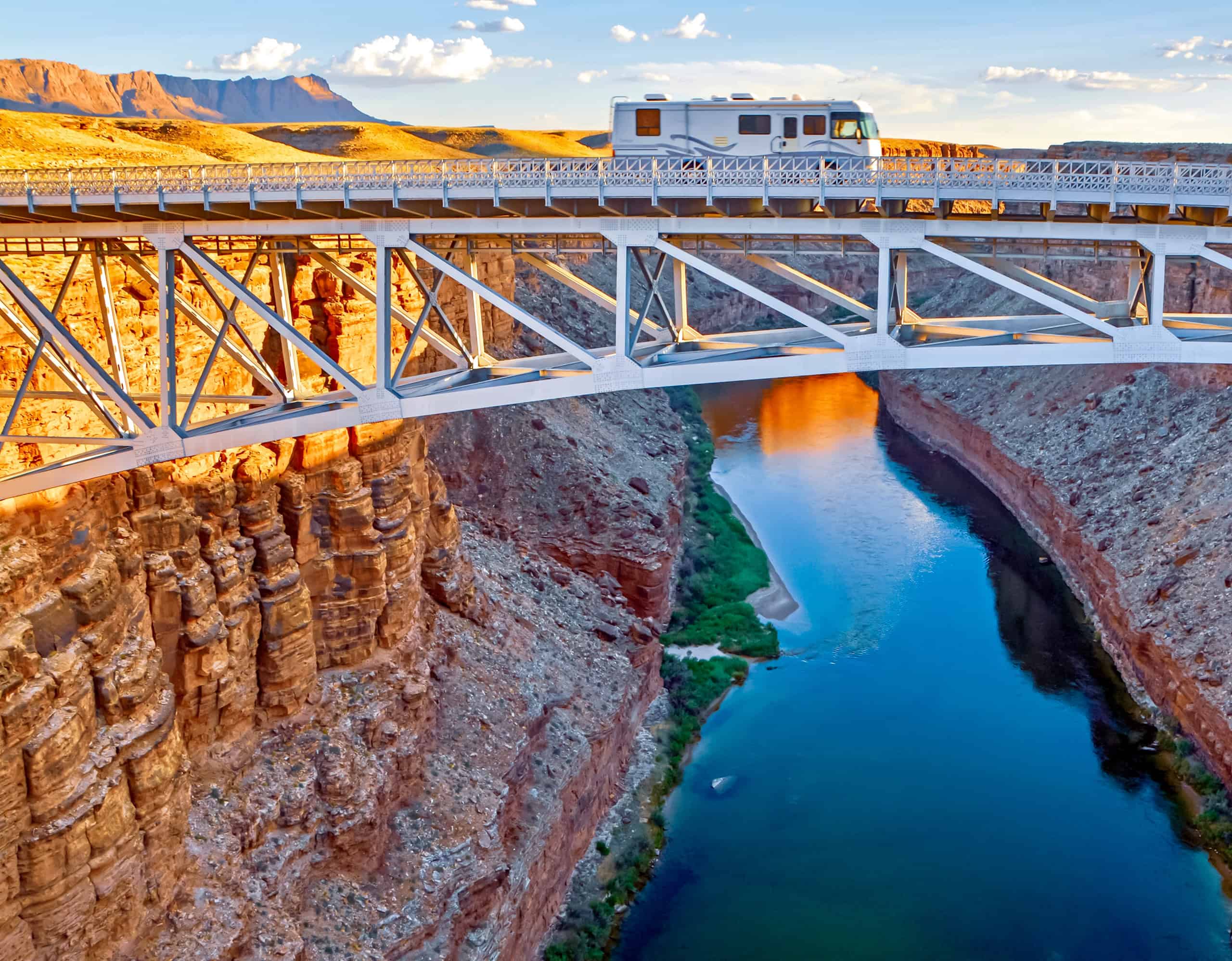 A Class A motorhome RV crossing a steel truss bridge high above the Colorado River cutting through a deep red rock canyon, with golden desert mesas and blue sky in the background