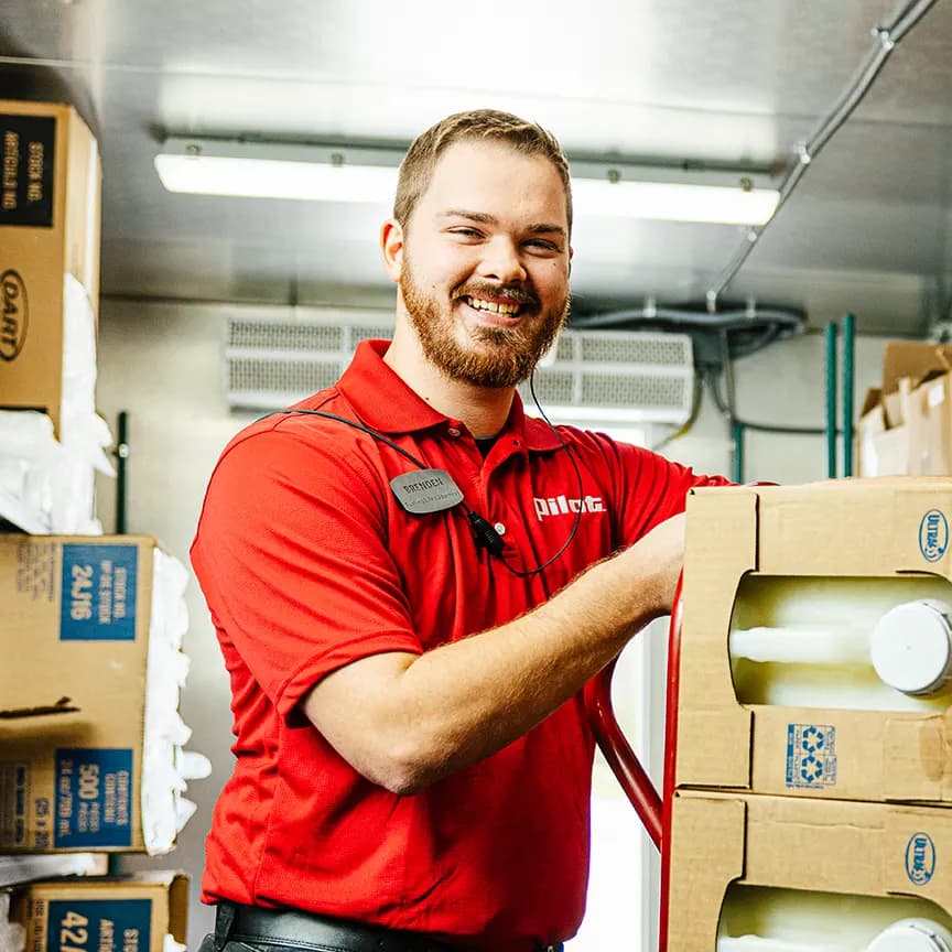 One Pilot Flying J employee, Brenden in a red polo, smiling inside a Pilot travel center store with storage shelves visible in the background