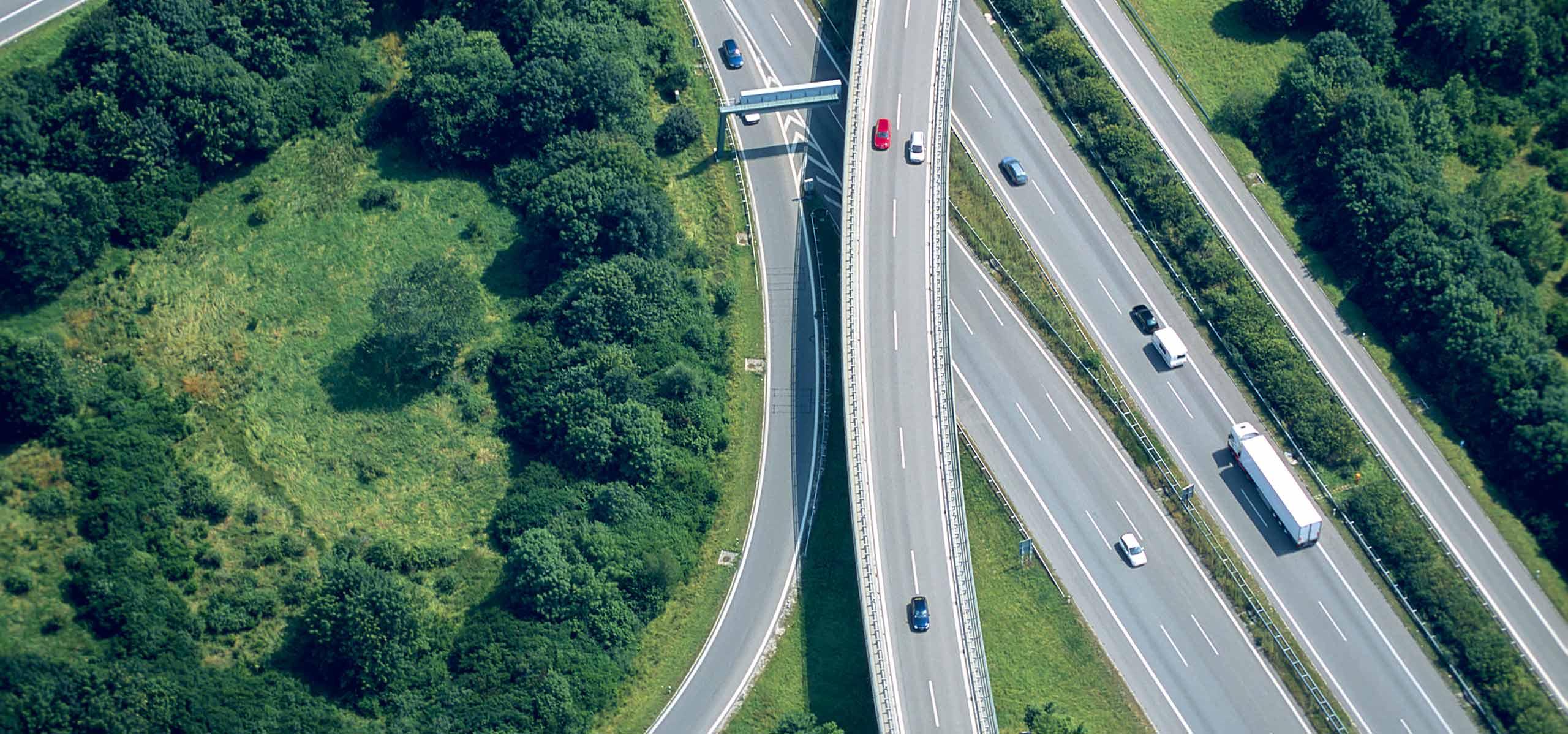 Aerial view of a highway on-ramp merging into a multi-lane motorway bordered by green trees, with cars and a semi-truck in traffic