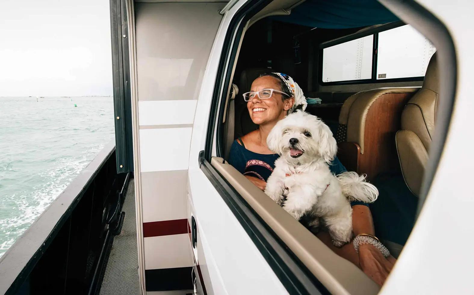 Smiling woman wearing white glasses and a floral headscarf holding a fluffy white Maltese dog while seated inside what appears to be an RV or motorhome cabin, with choppy ocean water visible through the open side door