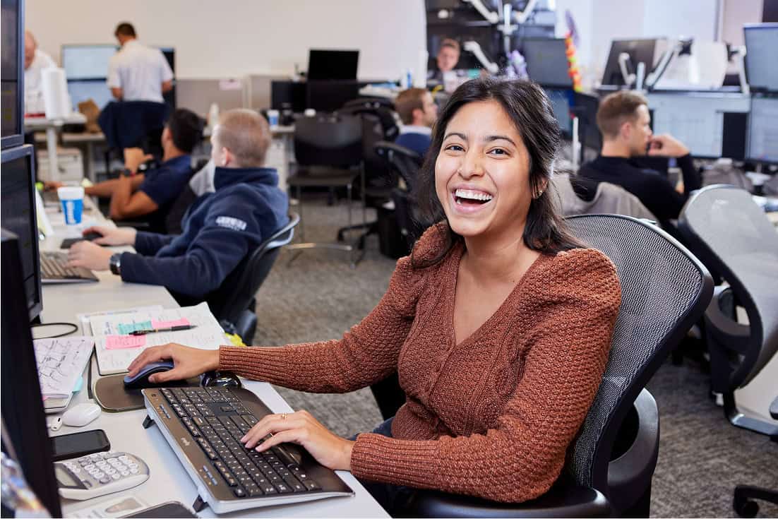 A smiling woman in a rust-colored sweater sitting at a desk with a keyboard and mouse, laughing toward the camera in a busy open-plan office with several colleagues working at computers in the background