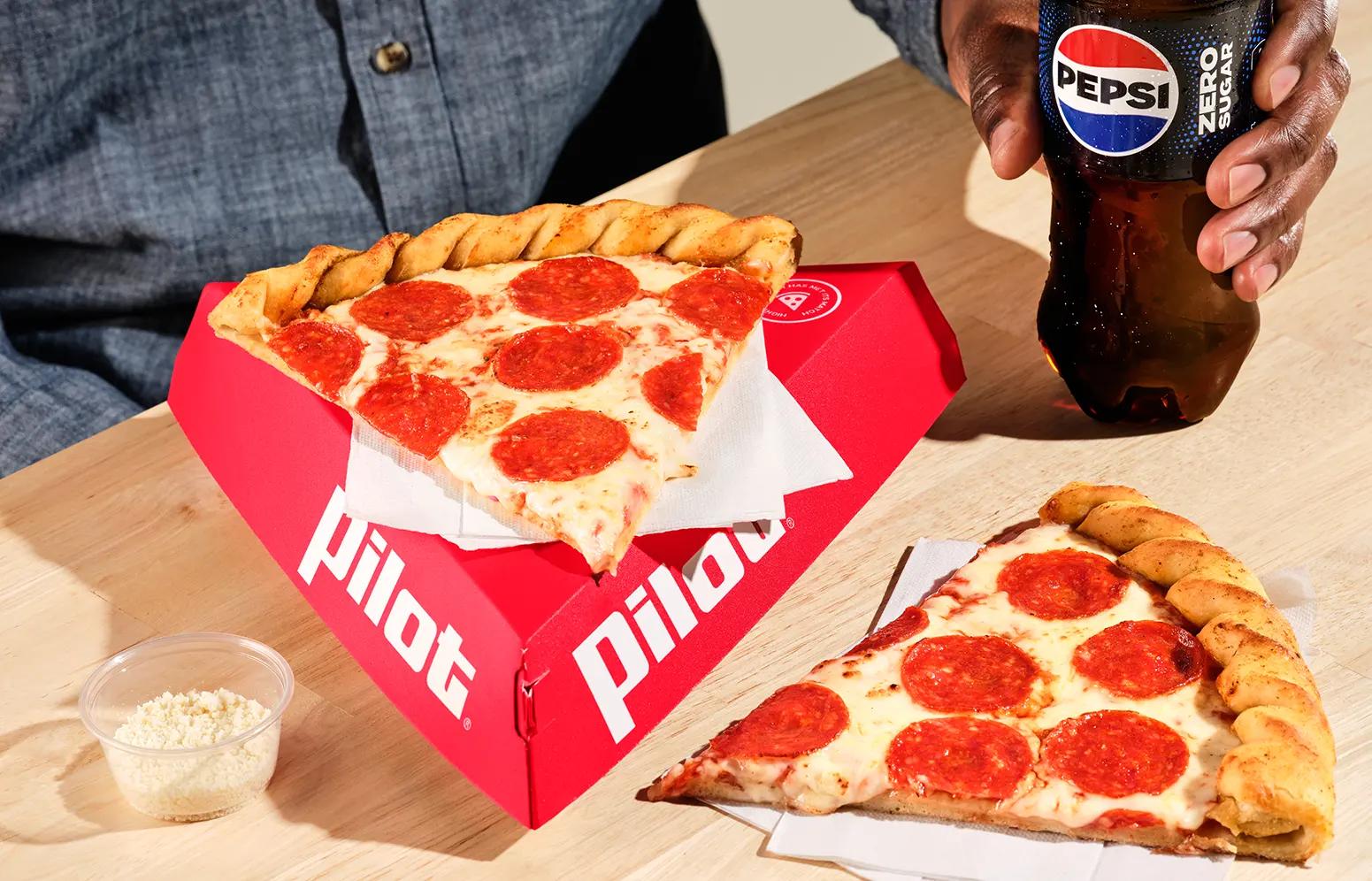 Two large pepperoni pizza slices with twisted crust from Pilot, one served in a red Pilot-branded triangular box, accompanied by a small cup of grated Parmesan cheese and a Pepsi Zero Sugar bottle held by a hand, all on a wooden table