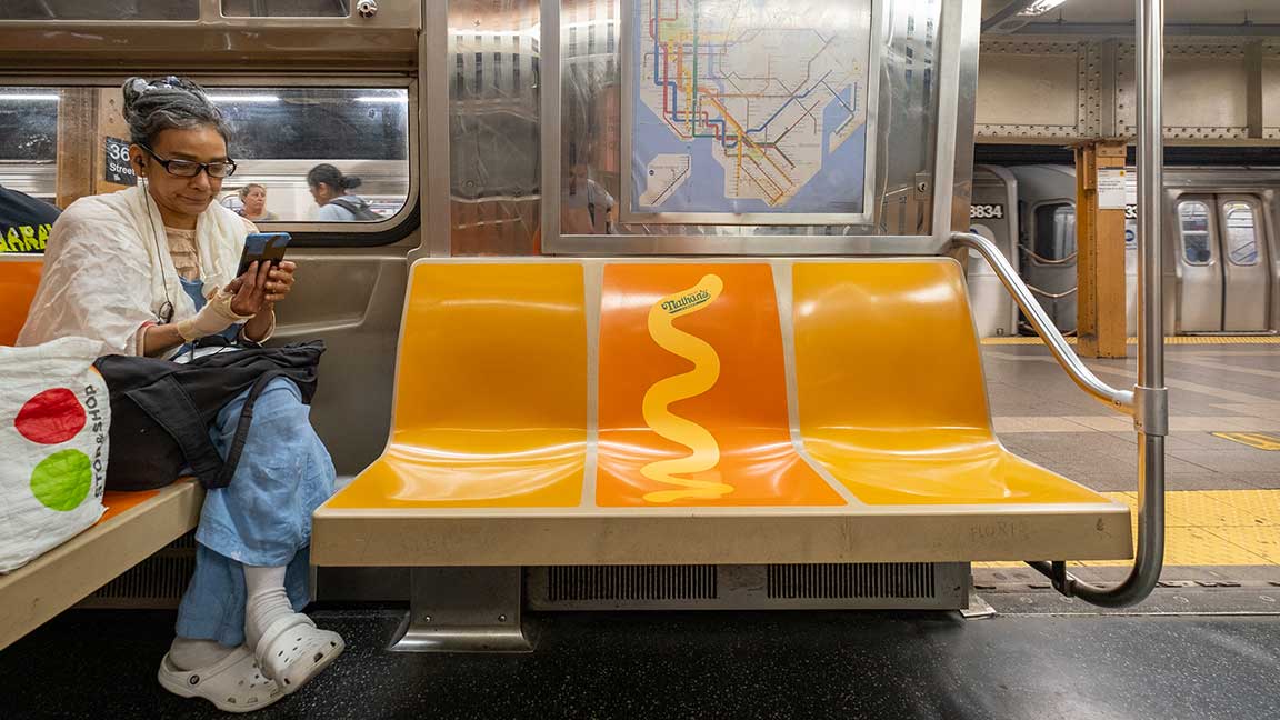 Nathan’s Famous NYC subway ad with a mustard decal on an orange seat in between two yellow seats on the N train, representing a hot dog in a bun  