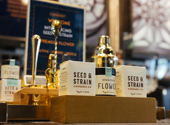 A premium retail display featuring Seed & Strain Cannabis Co. products. The packaging, labeled '1/8 OZ FLOWER,' is elegantly arranged on a wooden stand, accompanied by gold-toned accessories such as a cocktail shaker and stirring tools. The background features a sophisticated ambiance with warm lighting, decorative artwork, and a sign introducing the brand’s premium flower selections.