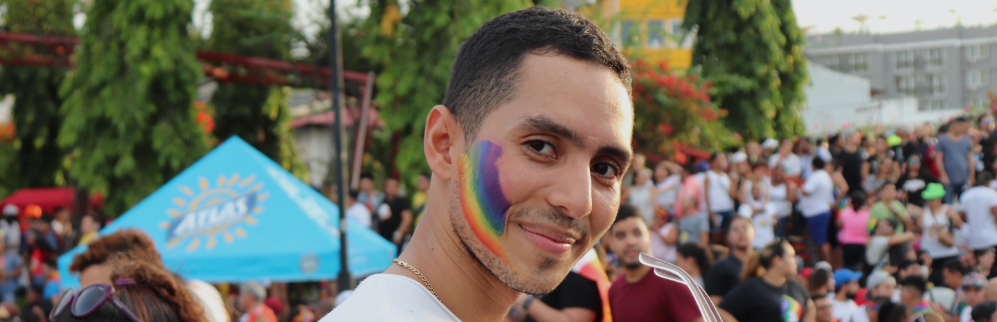 A young man with short dark hair and a light beard smiles at the camera during an outdoor Pride event. He has a vibrant rainbow painted on his cheek and is wearing a white t-shirt and a gold chain necklace. Behind him, a large crowd of people is gathered, many dressed in colorful attire. A bright blue tent with a sunburst logo is visible, along with green trees and urban buildings in the background. The atmosphere is lively and celebratory.