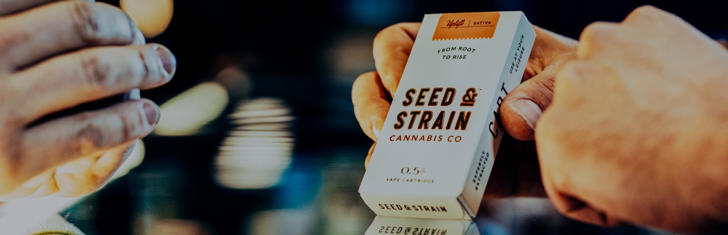 A close-up shot of two hands exchanging a Seed & Strain Cannabis Co. vape cartridge box. The packaging is white with bold brown text and features an orange label indicating 'Sativa' and the phrase 'From Root to Rise.' The background is blurred with warm lighting, creating a sleek and modern retail atmosphere.