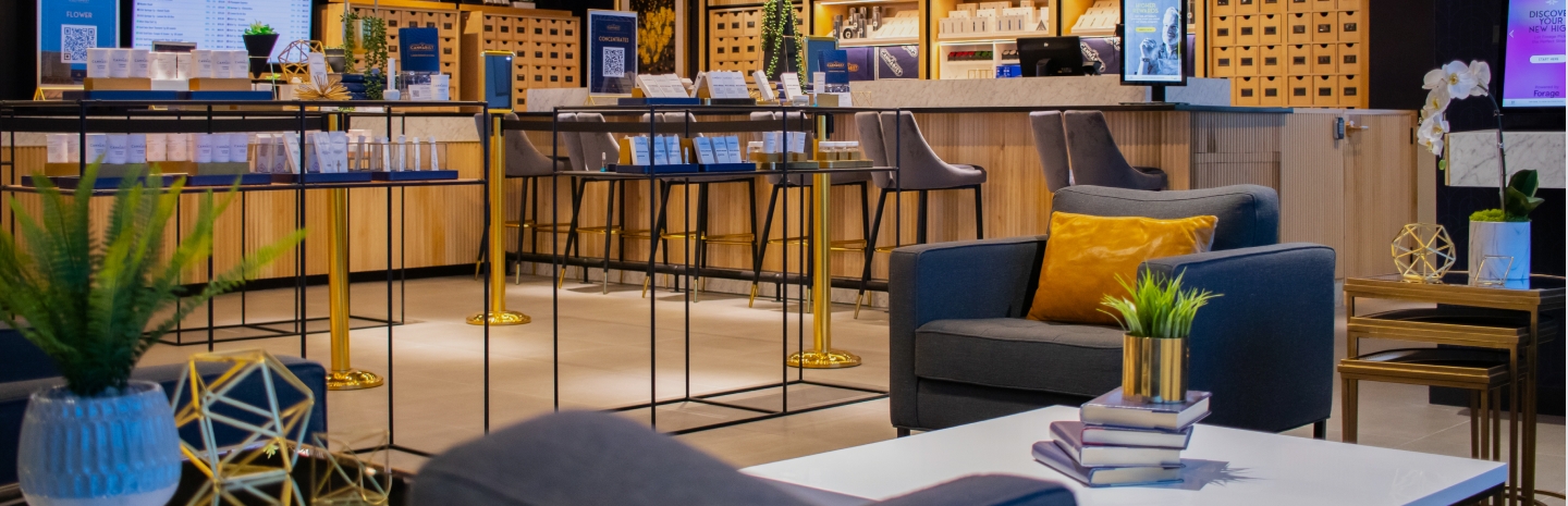 A Cannabist dispensary interior with a stylish, upscale design. The space features a lounge area with dark blue armchairs, gold-accented furniture, and a white coffee table adorned with a small stack of books and decorative geometric sculptures. A plush yellow pillow and green potted plants add warmth to the setting. In the background, the dispensary counter has high-top chairs with gold legs, sleek wooden cabinetry, and product displays on black metal-framed tables. Digital screens and signage provide product information. The ambiance is contemporary and inviting, blending luxury with a retail experience.