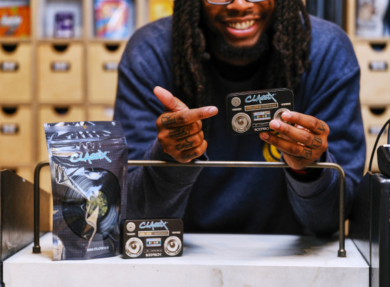 A smiling man with a navy blue sweatshirt holds a small, retro cassette tape-inspired cannabis product from Classix. He points at the product enthusiastically while standing behind a counter. In front of him are more Classix-branded products, including a sealed bag with a vinyl record design and a small matching container. The background features neatly arranged wooden drawers and shelves with colorful packaging.