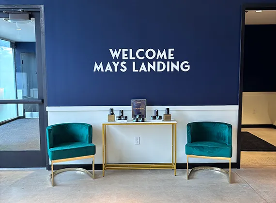 A welcoming lobby area at Cannabist Mays Landing, featuring a deep navy blue wall with bold white text reading 'WELCOME MAYS LANDING.' The space is elegantly furnished with two plush teal velvet chairs with gold accents, symmetrically placed on either side of a sleek gold-framed console table with a white top. The table displays a selection of cannabis-related products in stylish black and gold packaging. The modern and inviting atmosphere is complemented by natural light streaming in from a glass door on the left.