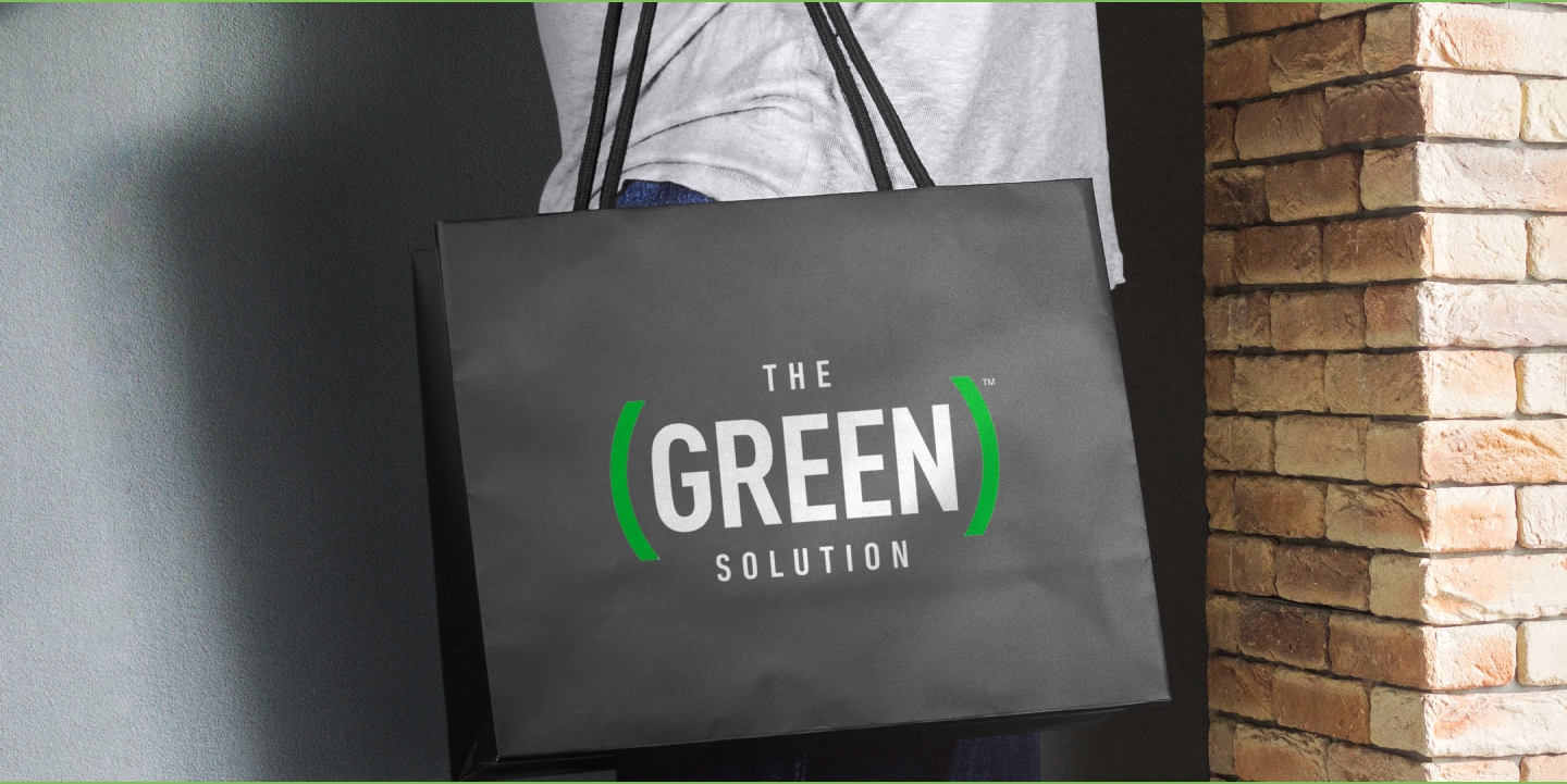 A person carrying a sleek black shopping bag with "The Green Solution" logo prominently displayed in white and green. The bold, modern typography stands out against the dark background. The person is dressed casually in a white shirt, and a brick wall is visible in the background.