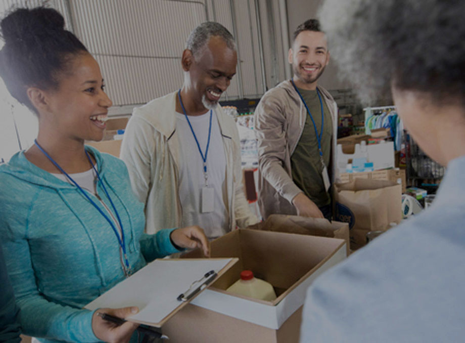 A diverse group of smiling volunteers wearing blue lanyards and ID badges, working together at a food donation center. One volunteer holds a clipboard while others pack essential goods, including a carton of milk, into boxes. The scene takes place inside a warehouse-like space filled with supplies, conveying a sense of teamwork and community service.