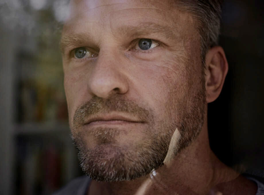 Close-up of a middle-aged man with short, graying hair and a beard, gazing thoughtfully out of a window. His blue eyes reflect the light, and his expression appears contemplative. The glass creates a subtle reflection on his face, adding depth to the image.