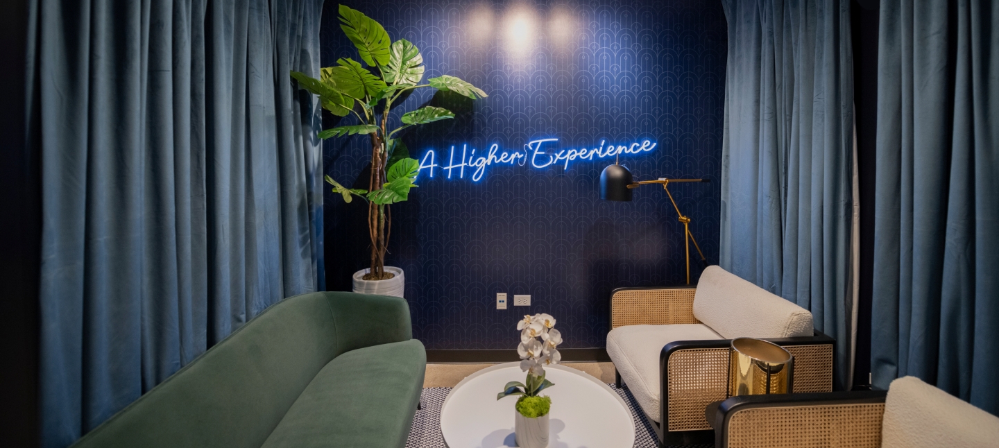 A stylish lounge area featuring a deep blue accent wall with a neon sign that reads 'A Higher Experience.' The space is furnished with a green velvet sofa, two rattan and white fabric armchairs, and a round white coffee table adorned with a small orchid plant. A tall potted plant stands in the corner, and a modern gold floor lamp with a black shade provides additional lighting. Plush blue curtains frame the space, creating a cozy and sophisticated atmosphere.