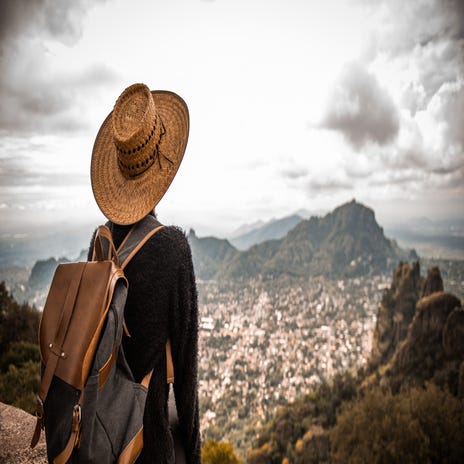 Man with a backpack and a hat sitting on a cliff and watching a city in the valley