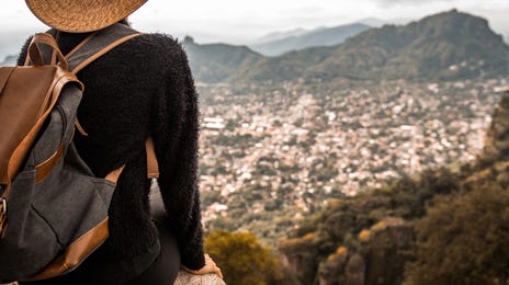 Man with a backpack and a hat sitting on a cliff and watching a city in the valley