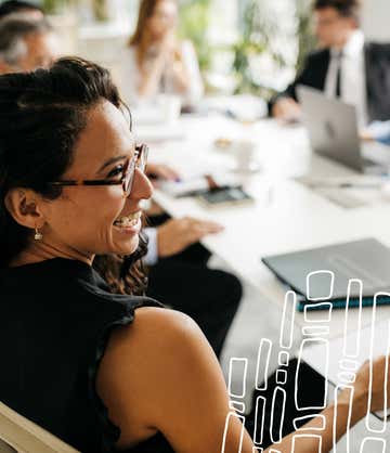 The board room table is full of people working together as a woman smiles at the person on her right.