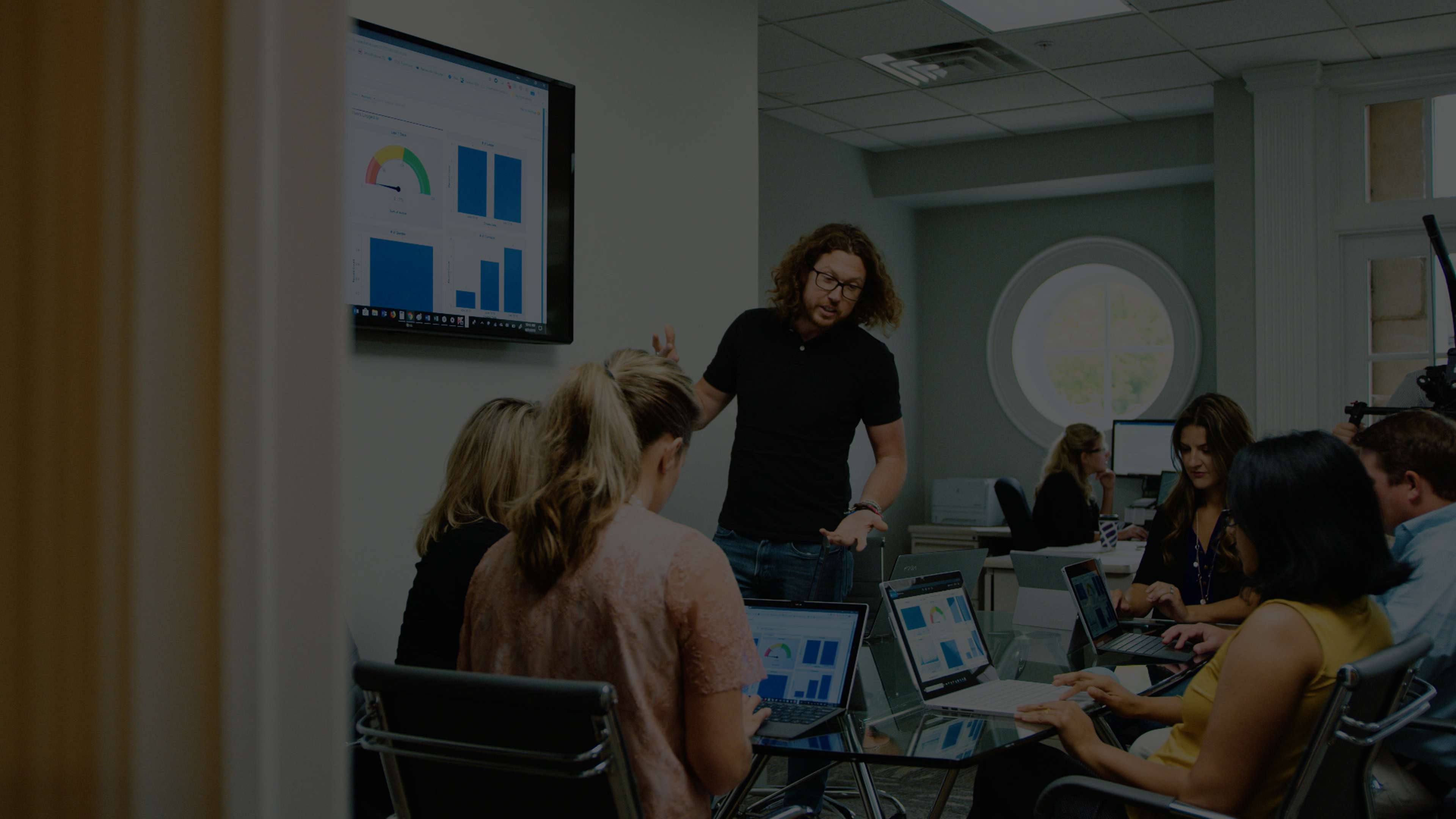A business meeting is taking place. An employee stands near a large screen sharing graphs with colleagues seated around a table with laptops.