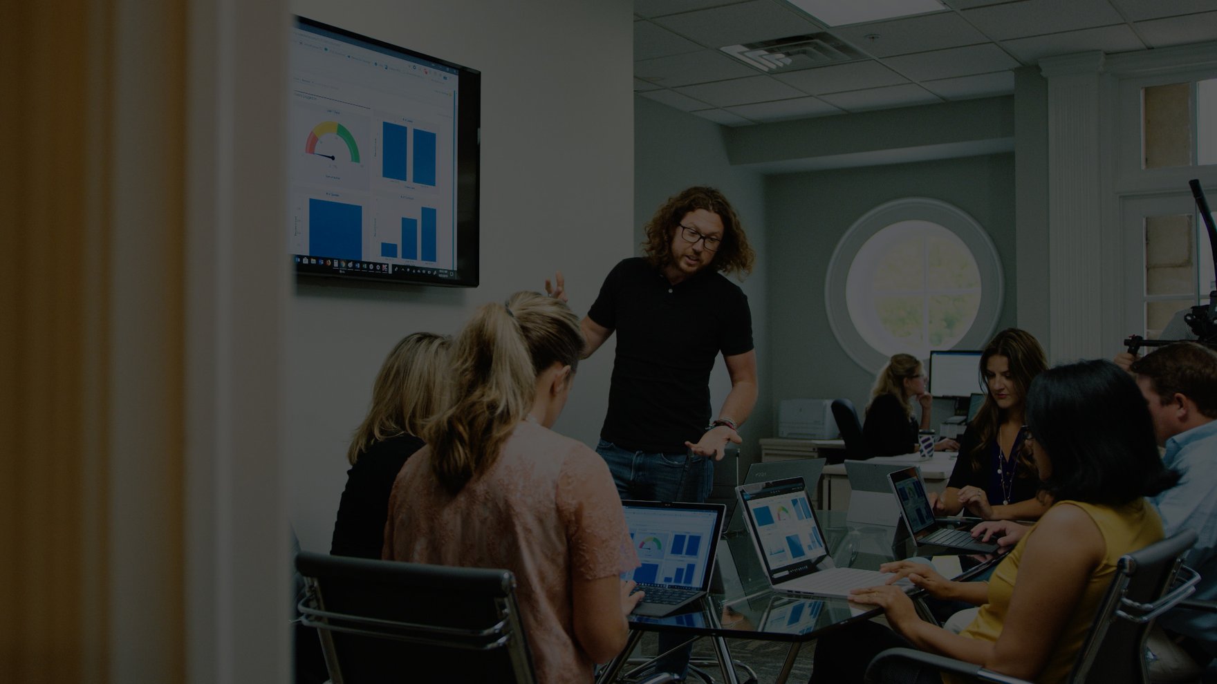 A business meeting is taking place. An employee stands near a large screen sharing graphs with colleagues seated around a table with laptops.