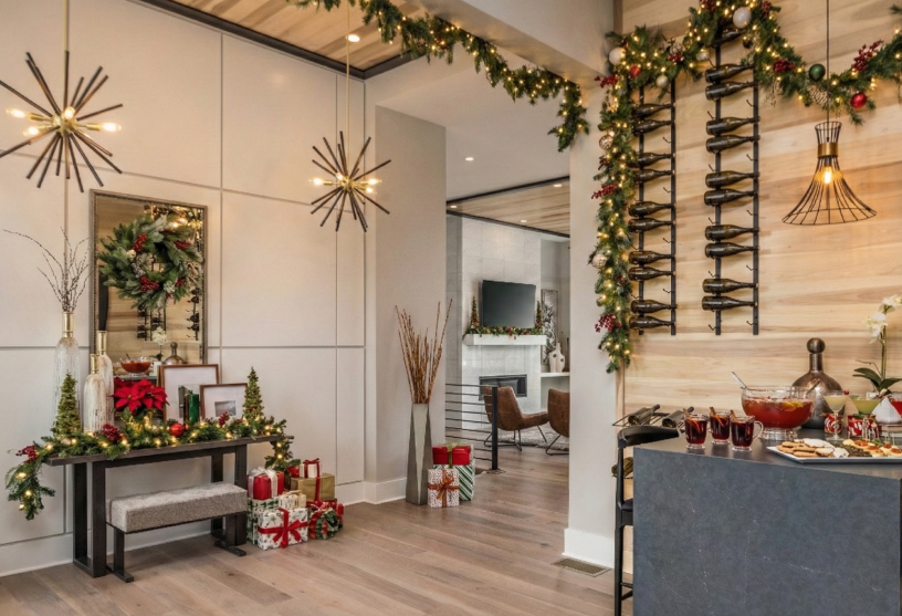 Festive interior with garlands, modern chandeliers, and a bar setup. Wrapped gifts and poinsettias adorn a table, creating a warm, holiday ambiance.