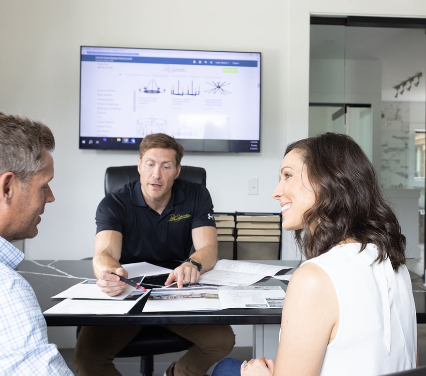A consultant guides a couple through new house plans. The trio reviews blueprints on a desk, a monitor displaying product details in the background, in a bright modern office.