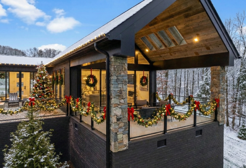 Modern house deck decorated with Christmas garlands, wreaths, and lights, surrounded by snowy trees under a partly cloudy sky.