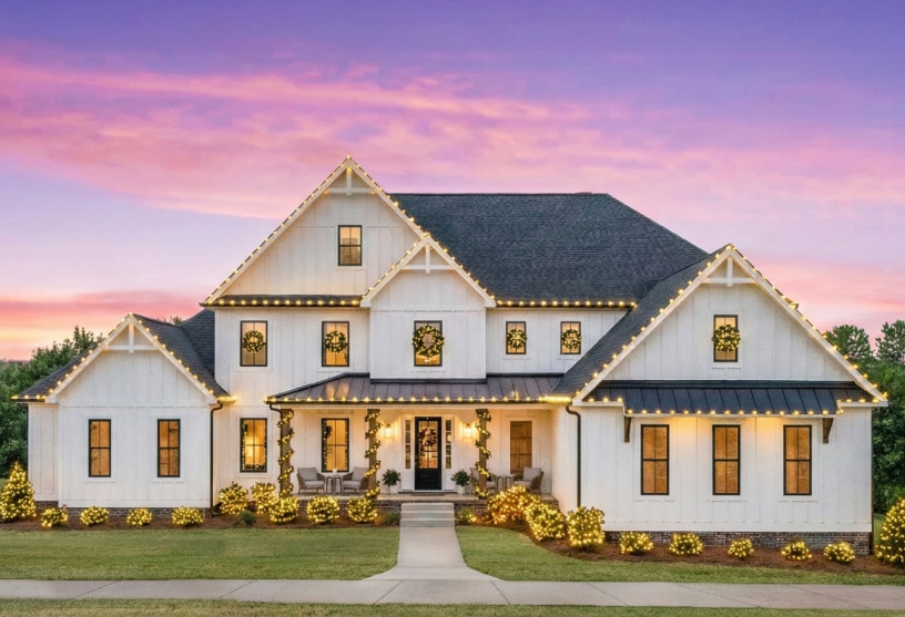 A large farmhouse with white siding is decorated with warm white string lights, surrounded by lit shrubs. The sky is a vibrant purple and pink at sunset.